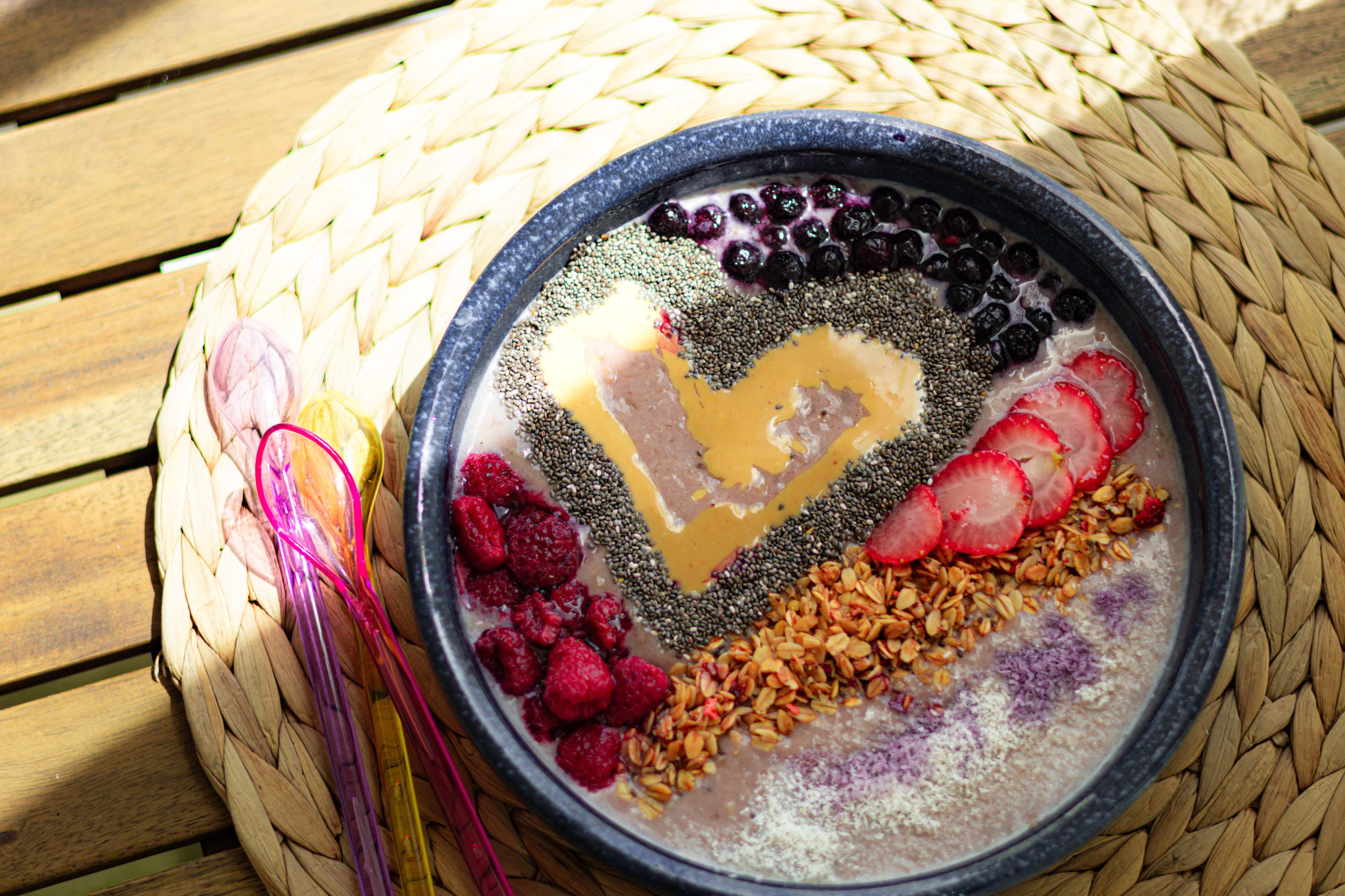 A smoothie bowl topped with a peanut butter heart, chia seeds, blueberries, strawberries, raspberries, and granola, set on a woven placemat with colorful spoons