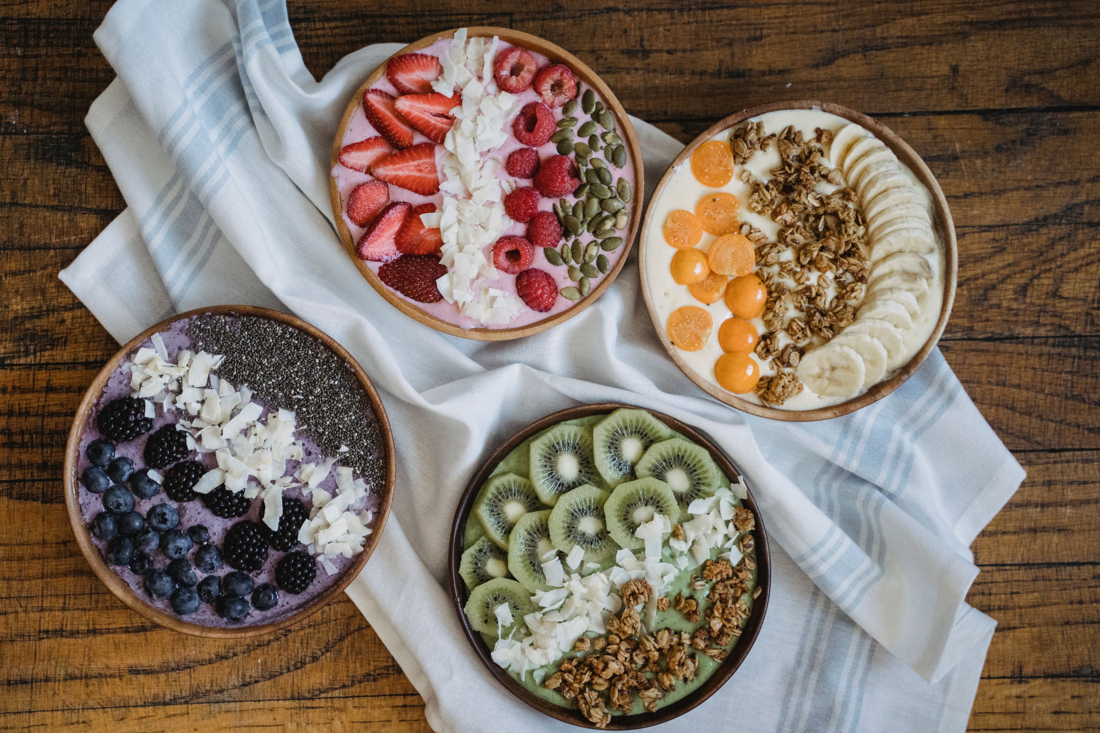 Four colorful smoothie bowls arranged overhead on a wood table with a striped linen cloth — a pink berry bowl with strawberries and pumpkin seeds, a purple blueberry-blackberry bowl with chia, a green kiwi bowl with granola, and a golden bowl with gooseberries, granola, and banana