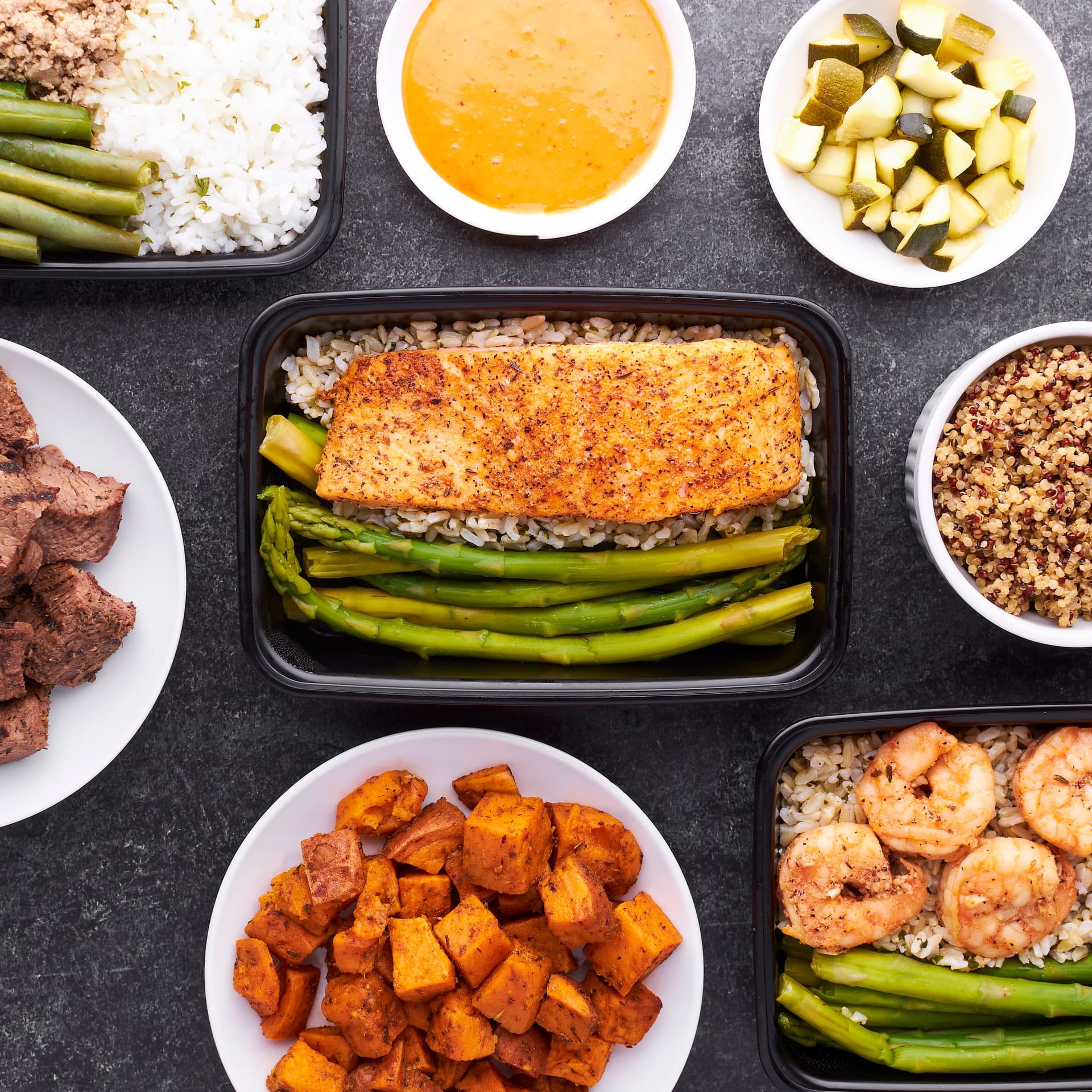An overhead meal-prep spread on dark slate — seasoned salmon with rice and asparagus at the center, surrounded by steak bites, roasted sweet potatoes, quinoa, shrimp with asparagus, and zucchini